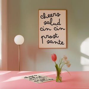 May include: A rectangular wall print in a thin wooden frame featuring the black handwritten text cheers, salud, cin cin, prost, and sante. A matching print lies on a pink surface next to a clear glass vase containing pink and white tulips.