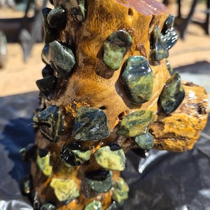 May include: A cylindrical piece of polished light brown wood decorated with numerous embedded dark green and yellow mottled stones. The stones are irregularly shaped and have a glossy finish, contrasting with the textured grain of the wood surface.