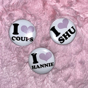 Puede incluir: Tres pines de botón blancos con texto negro y un corazón rosa. Los pines dicen "Me encanta Coups", "Me encanta Shu" y "Me encanta Hannie".