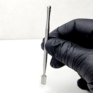 May include: A silver metal tool with a rounded tip and a flat, rectangular blade. The tool is held in a gloved hand.