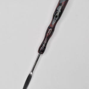May include: A palette knife with a decorative handle. The handle is a mix of red, black, and gray with a skull design. The blade is a flat, silver color.