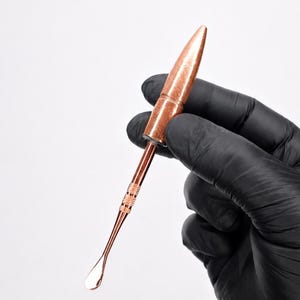 Puede incluir: Una herramienta de metal color cobre con un mango en forma de bala y un vástago delgado y alargado que termina en una pequeña cuchara plana y redondeada. El mango tiene una sección de agarre texturizada con un patrón entrecruzado.