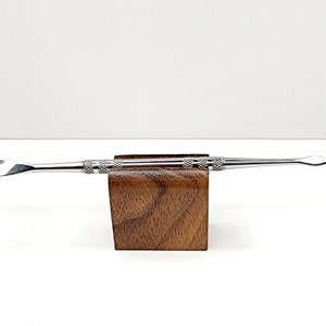 May include: A silver metal dab tool with a rounded tip and a textured handle. The tool is displayed on a small wooden stand.