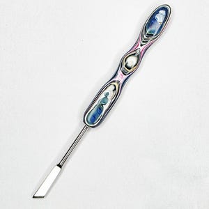 Puede incluir: Una herramienta de metal con una hoja larga, delgada y de color plateado unida a un mango contorneado. El mango muestra un patrón de capas multicolor con remolinos de azul, blanco, negro y rosa, creando una apariencia suave, pulida y orgánica.