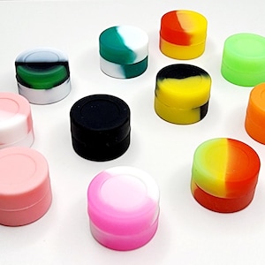 Op de afbeelding: Een verzameling kleine, ronde siliconen containers in verschillende kleuren, waaronder roze, groen, oranje, geel en zwart. Sommige containers hebben een effen kleur, terwijl andere een tweekleurig ontwerp hebben. De containers zijn gerangschikt op een wit oppervlak.