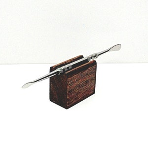May include: A silver metal tool with a rounded tip and a textured handle is displayed on a dark brown wooden stand.