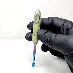 Puede incluir: Una herramienta de metal iridiscente multicolor con una punta puntiaguda y un borde plano redondeado. La herramienta tiene un mango marmoleado verde, morado y blanco.