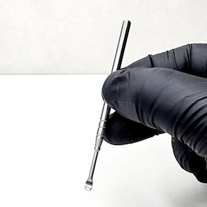 Könnte beinhalten: Ein silbernes Dab-Werkzeug aus Metall mit abgerundeter Spitze. Das Werkzeug wird in einer behandschuhten Hand gehalten.