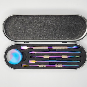 Puede incluir: Una caja de metal plateada con un interior de espuma negra contiene un juego de cuatro herramientas de metal iridiscente y un pequeño recipiente de vidrio azul y morado.