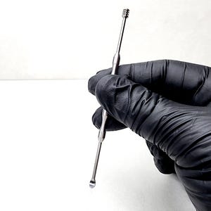 Puede incluir: Una herramienta de metal plateado con una punta redondeada y una espiral en la parte superior. La herramienta se sostiene en una mano enguantada de negro.