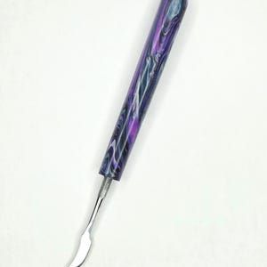 May include: A cuticle pusher tool featuring a cylindrical handle with a marbled pattern in shades of purple, black, and white. The handle connects to a polished silver-colored metal tip that is curved and tapered to a rounded point at the end.