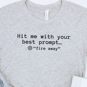 May include: Gray t-shirt with the text "Hit me with your best prompt... "fire away" printed on it.