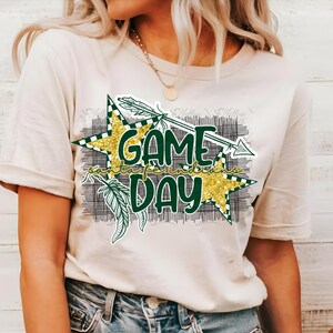 May include: A white t-shirt with a green and gold graphic design that says "Game Day". The graphic features a star, an arrow, and feathers.