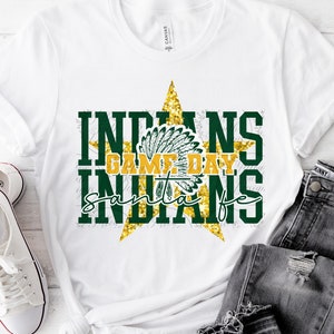 May include: White t-shirt with a gold glitter star design. The star has a green and gold headdress inside. The text "Indians Game Day Santa Fe" is written in green and gold letters.
