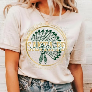 May include: Beige t-shirt featuring a gold and green graphic of a Native American headdress and the words "Santa Fe Indians" in a gold font. The design is set within a gold circle with a glitter effect.