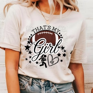 Puede incluir: Una camiseta blanca con el texto "That's My Girl" en letras negras. El texto está rodeado de un balón de fútbol negro, estrellas negras y una silueta negra de una atleta femenina corriendo con un corazón que tiene el número 8 dentro.