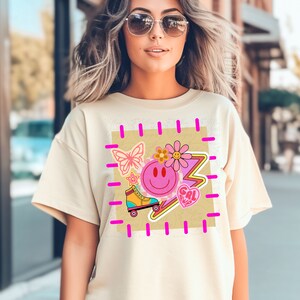 Peut inclure: Un t-shirt beige avec un imprimé graphique d'un visage souriant, d'un papillon, d'une fleur, d'un patin à roulettes, d'un éclair et du texte "GAL PWR".