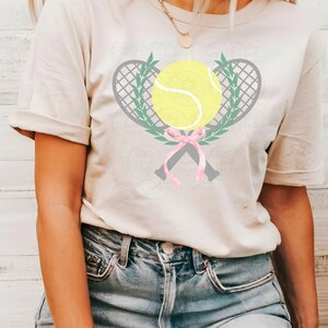 Puede incluir: Una camiseta blanca con un dise&ntilde;o de raqueta de tenis y pelota grises. Las raquetas est&aacute;n cruzadas y atadas con una cinta rosa. Las hojas verdes rodean la pelota y las raquetas.