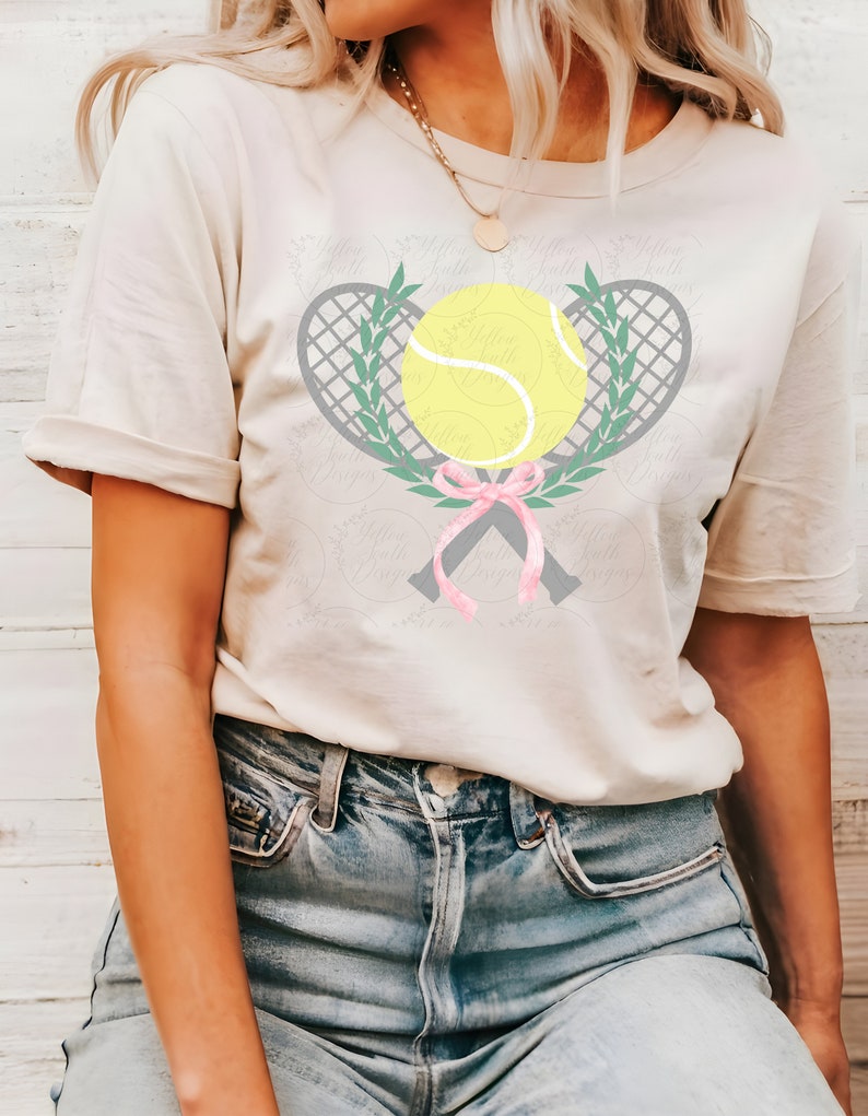 Puede incluir: Una camiseta blanca con un dise&ntilde;o de raqueta de tenis y pelota grises. Las raquetas est&aacute;n cruzadas y atadas con una cinta rosa. Las hojas verdes rodean la pelota y las raquetas.