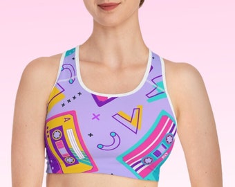May include: A lilac sports bra with a white trim and a colorful 80s-themed design. The pattern includes cassette tapes, geometric shapes, and abstract designs in yellow, pink, teal, and purple.