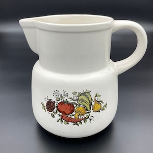 May include: White ceramic jug with a handle and a colourful vegetable design. The design features a red tomato, green pepper, yellow onion, brown mushroom, and orange carrot.
