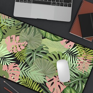 Puede incluir: Una alfombrilla de rat&oacute;n negra con un dise&ntilde;o de hojas tropicales. El dise&ntilde;o presenta hojas verdes y rosas sobre un fondo negro.