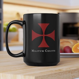 May include: Black ceramic mug featuring a red cross design with the words "Militum Christe" in white. The mug has a large handle and is likely used for hot beverages. The design is centered on the mug's surface.