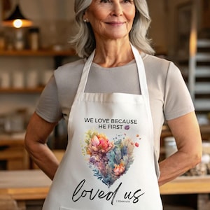 Op de afbeelding: Witte schort met een hartvormig bloemmotief in roze, paarse en blauwe tinten. De schort heeft de tekst "WE LOVE BECAUSE HE FIRST" en "loved us" met een bijbelversverwijzing.