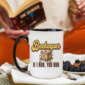 May include: White ceramic mug with a black handle and rim. The mug features the text "Beekeeper" in a retro font, along with an illustration of a beehive and the phrase "IF I RUN, YOU RUN". A plate with blueberries and a cookie is also visible.
