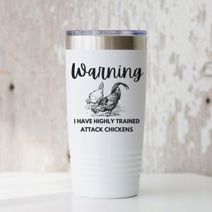May include: White travel mug with a silver lid. The mug features a black and white illustration of a rooster and hen, with the text "Warning I have highly trained attack chickens."