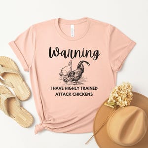 Könnte beinhalten: Hellrosa T-Shirt mit dem Text "Warning I Have Highly Trained Attack Chickens" und einer schwarzen Illustration eines Hahns und einer Henne. Das Shirt ist mit beigen Sandalen und einem braunen Hut gestylt.