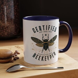 May include: White ceramic mug with a navy blue interior and handle. The mug features a detailed illustration of a bee with the words "CERTIFIED RAW LOCAL WILD HANDCRAFTED HONEY BEEKEEPER" in a vintage style. A spoon and a macaron are on a wooden surface.