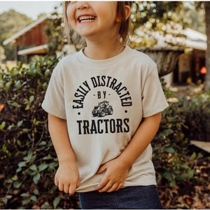 May include: A beige t-shirt with the text "EASILY DISTRACTED BY TRACTORS" in a black, circular design. The design includes a tractor graphic. The shirt is worn by a child.
