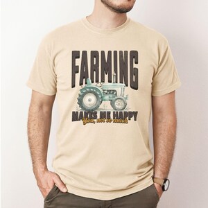 May include: Beige t-shirt with the text "FARMING" above a light blue tractor graphic. Below the tractor, the text reads "MAKES ME HAPPY" with "You, not so much" in a smaller font. The shirt has short sleeves.
