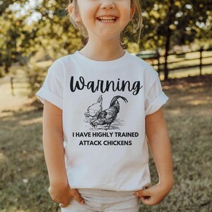 Puede incluir: Camiseta blanca con la frase "Warning" sobre una ilustración de dos gallinas. Debajo de la ilustración, el texto dice "I have highly trained attack chickens."