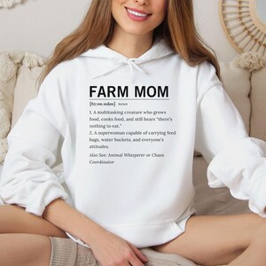May include: A white hoodie featuring the text "FARM MOM" in black, accompanied by a definition. The definition describes a multitasking individual involved in food production, cooking, and farm duties. The hoodie is a casual garment.
