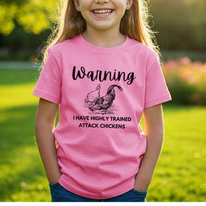 Puede incluir: Camiseta rosa con el texto "Warning I have highly trained attack chickens" y una ilustración en blanco y negro de dos gallinas. La camiseta la lleva una persona.