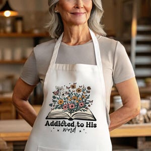May include: White apron with a floral design and the text "Addicted to His word." The design features an open book with flowers and butterflies. The apron is worn by a person.