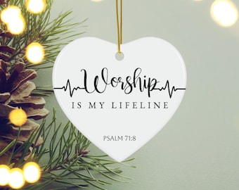 Puede incluir: Un adorno blanco en forma de corazón con las palabras "Worship is my Lifeline" en escritura negra, acompañado de una línea de latido cardíaco. El texto "Salmo 71:8" está debajo. Se adjunta una cuerda dorada para colgar. El fondo incluye vegetación y luces suaves.