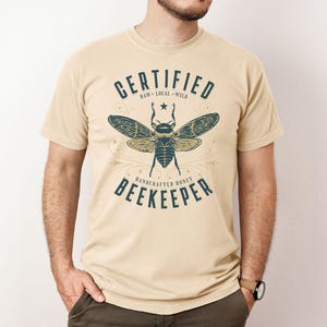 May include: Beige t-shirt featuring a navy blue bee graphic and the text "CERTIFIED BEEKEEPER". The design also includes the words "RAW LOCAL WILD" and "HANDCRAFTED HONEY". The shirt is short-sleeved and worn by a person.