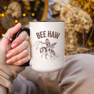 May include: White ceramic mug with a black handle and rim, featuring the text "BEE HAW" above an illustration of a beekeeper riding a bee. The design is in brown. The mug is held by a person.