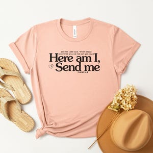 May include: A light pink t-shirt with the text "Here am I, Send me" in black. The shirt is styled with a pair of woven sandals and a brown hat with a flower. The text is from Isaiah 6:8.