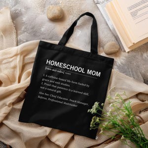Könnte beinhalten: Schwarze Tragetasche mit den weißen Worten "HOMESCHOOL MOM". Die Definition von "Homeschool Mom" ist darunter aufgeführt. Die Tasche hat schwarze Griffe und liegt auf einer neutralfarbenen Oberfläche.