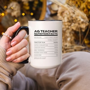 Può includere: Tazza in ceramica bianca con manico e bordo neri, con la scritta "AG TEACHER NUTRITION FACTS" e un elenco umoristico dei valori giornalieri. La tazza è tenuta da una persona con un braccialetto di perle.