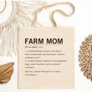 May include: Cream-colored tote bag with the text "FARM MOM" in black. The definition of "Farm Mom" is printed below the title. The bag has two handles and is surrounded by decorative items.