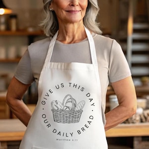 Op de afbeelding: Witte schort met een zwarte illustratie van een broodmand. De tekst "GIVE US THIS DAY OUR DAILY BREAD" omcirkelt de mand, met "Matthew 6:11" eronder. De schort wordt door een persoon gedragen.