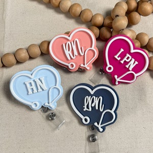 May include: Four heart-shaped badge holders with a stethoscope design. The holders are in pink, blue, red, and navy blue with white text that reads "RN", "RN", "LPN", and "LPN".