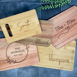 May include: Five different wooden cutting boards with personalized engravings. The boards are made of bamboo and acacia wood and feature various fonts and designs. The engravings include "Smith Family", "The Howards Est. 2020", "The Daniels", "Thompson", and "Tina + Mike".