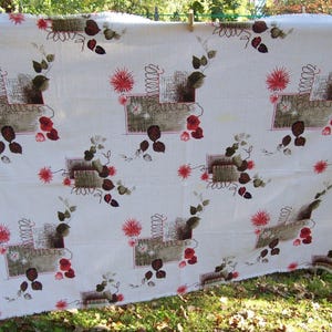 May include: White fabric with a repeating pattern of stylized floral and geometric elements. The design includes brown, red, and green accents, with a textured appearance. The fabric is hanging outdoors, suggesting it's a tablecloth or similar textile.