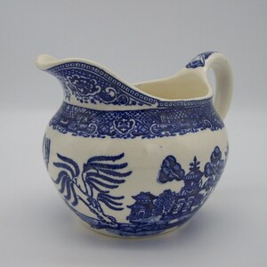 May include: A white ceramic creamer with a blue and white willow pattern. The creamer has a rounded body and a spout on one side. The handle is on the opposite side of the spout.